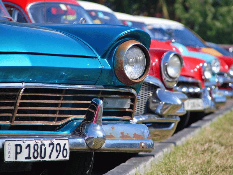 Nahaufnahme eines türkisfarbenen Oldtimers mit rostiger Stoßstange und kubanischem Kennzeichen vor einer Reihe bunter klassischer Autos in Havanna, Kuba