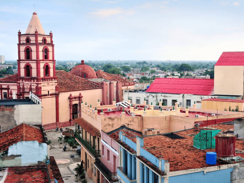 Luftaufnahme einer Stadt mit markantem rosa-roten Kirchturm, Kuppelgebäude und zahlreichen Terrakottadächern in Trinidad, Kuba