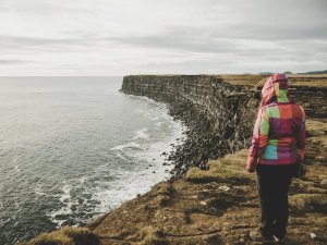 Person in bunter Jacke steht auf einer grasbewachsenen Klippe mit Blick auf das Meer in Island