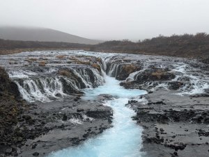 Leuchtend blaues Gletscherwasser fließt über dunkle Felsen und Wasserfälle unter einem nebligen Himmel in Island