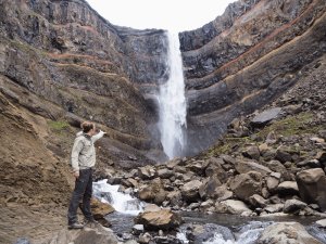 Person zeigt auf einen hohen Wasserfall, der über geschichtete Felsen in einen steinigen Fluss stürzt in Island