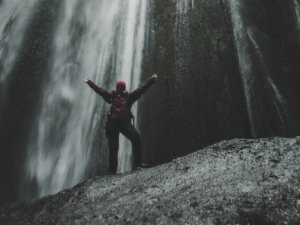 Person in roter Jacke und Mütze steht mit ausgestreckten Armen auf einem Felsen vor einem Wasserfall in Island