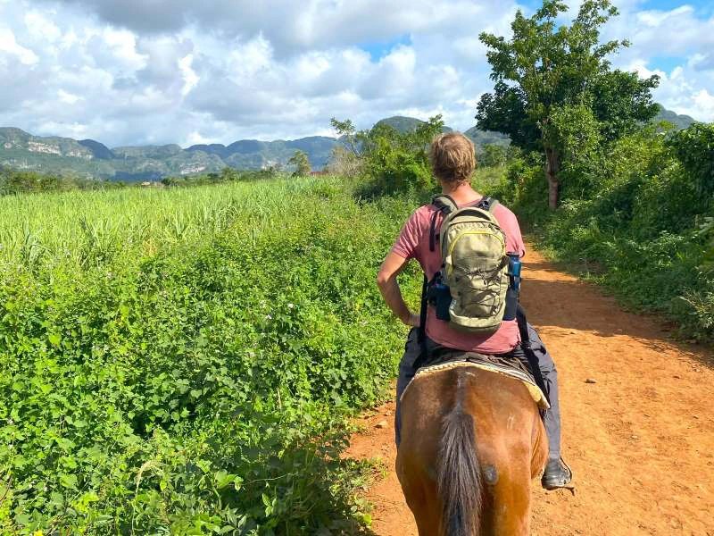 Person reitet auf einem Pferd über einen Sandweg durch üppig grüne Felder mit Bergen in der Ferne unter bewölktem Himmel in Viñales, Kuba