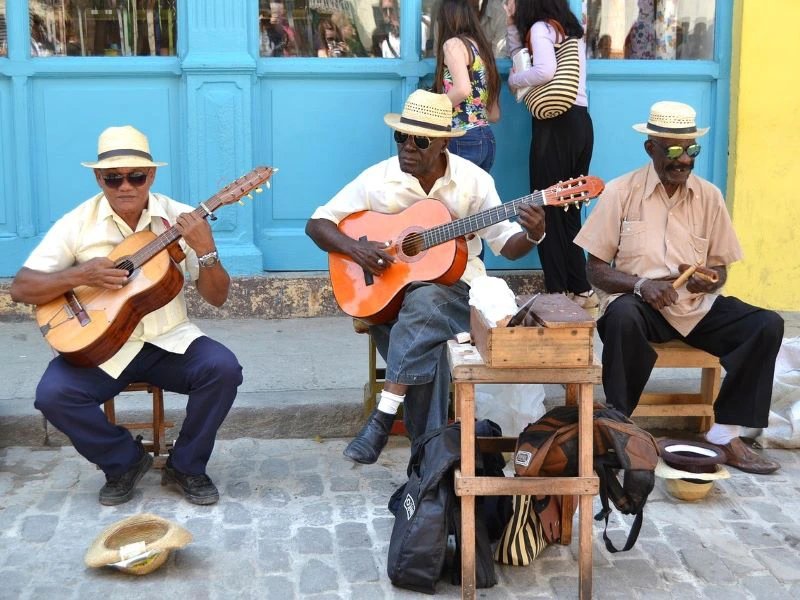 Drei Musiker mit Strohhüten und Sonnenbrillen spielen Gitarre und Percussion auf einer gepflasterten Straße in Trinidad, Kuba