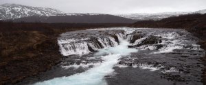 Leuchtend blauer Fluss fließt über dunkle Felsen zwischen brauner Vegetation und schneebedeckten Bergen unter grauem Himmel in Island