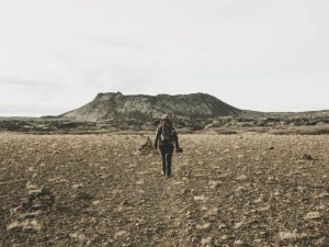 Einsamer Wanderer mit Rucksack läuft über eine karge felsige Ebene auf einen dunklen Hügel unter bewölktem Himmel zu in Island