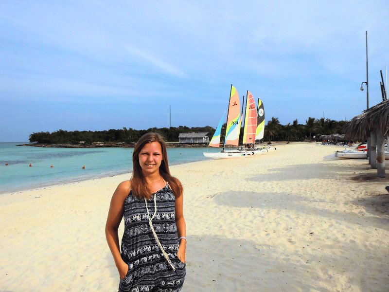Lächelnde junge Frau an einem Sandstrand mit türkisfarbenem Meer und bunten Segelbooten im Hintergrund in Cayo Santa María, Kuba