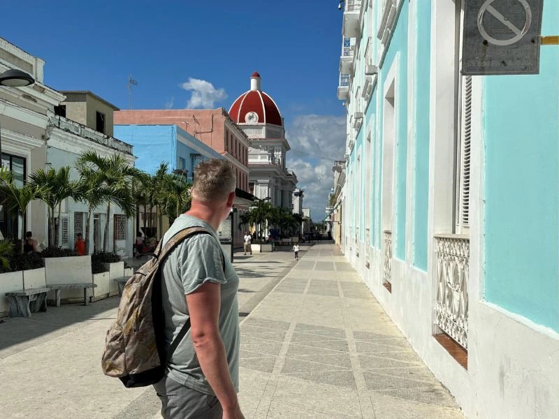 Mann mit Rucksack auf einer sonnigen Fußgängerzone mit bunten Pastellgebäuden und markantem roten Kuppelbau im Hintergrund in Cienfuegos, Kuba