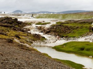 Bach fließt durch eine dampfende geothermische Landschaft mit weißen Mineralablagerungen, grünem Gras und badenden oder wandernden Menschen in Island