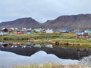 Bunte Häuser eines Dorfes spiegeln sich im ruhigen Wasser vor hohen Bergen unter bewölktem Himmel in Grönland