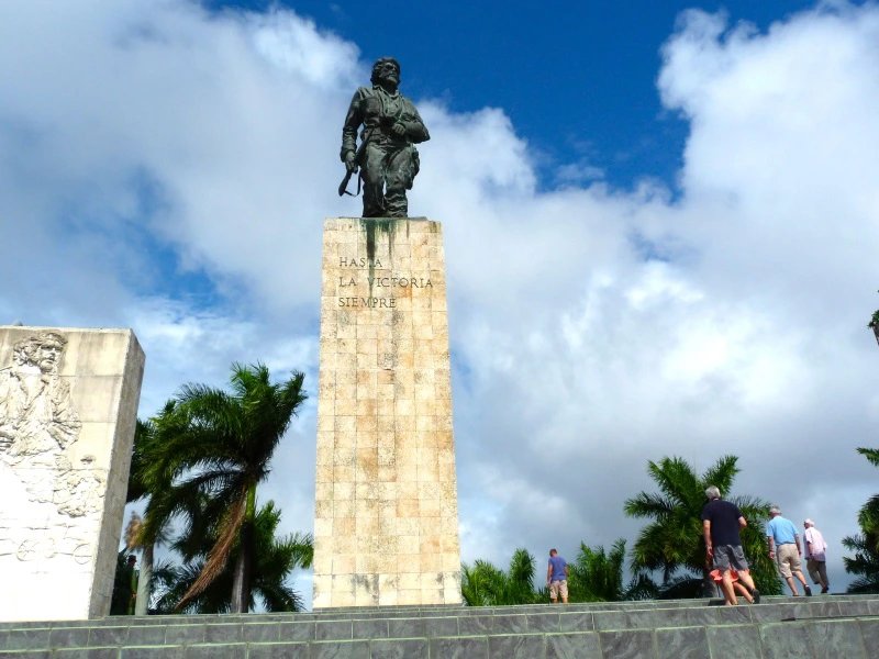 Bronzestatue von Che Guevara auf hohem Sockel mit der Inschrift „Hasta La Victoria Siempre“, mit Denkmälern, Palmen und Menschen unter bewölktem Himmel in Santa Clara, Kuba