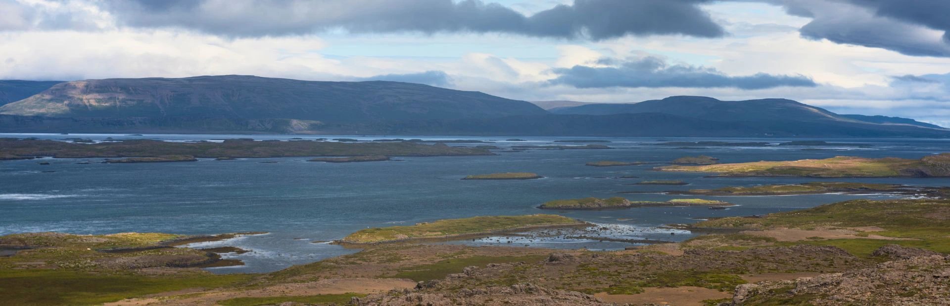 Panoramablick auf eine Bucht mit zahlreichen kleinen grünen Inseln und Bergen in der Ferne unter bewölktem Himmel in Island