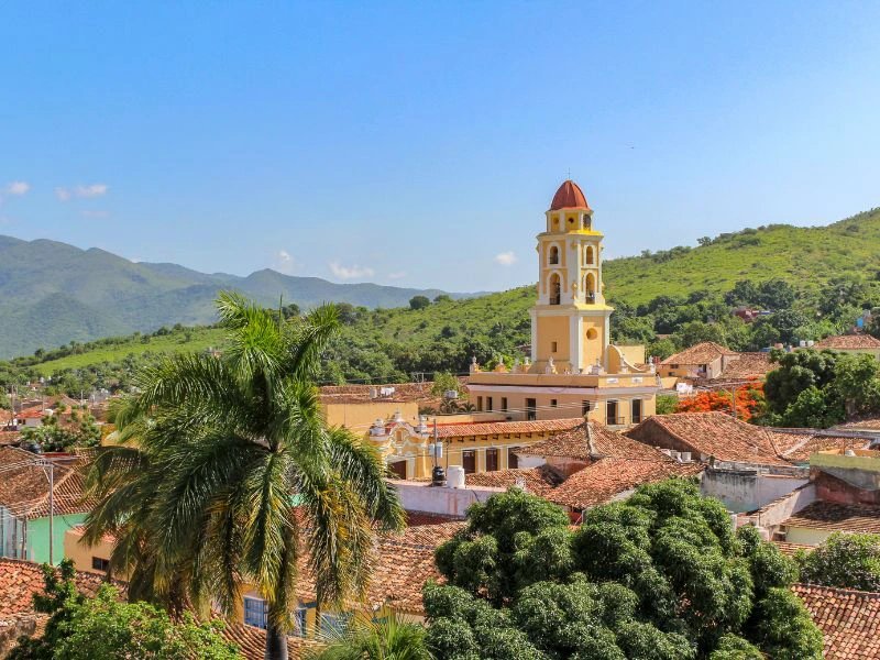 Panoramablick über eine Stadt mit gelbem Glockenturm, roten Ziegeldächern, großer Palme und grünen Bergen unter blauem Himmel in Trinidad, Kuba