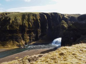 Fluss fließt durch eine tiefe moosbedeckte Schlucht entlang eines Wasserfalls unter einem Felsbogen mit Menschen am Ufer in Island