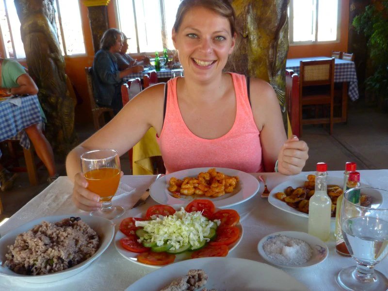 Lächelnde Frau an einem Restauranttisch mit Tellern voller Garnelen, Reis mit Bohnen, Salat und einem Glas Saft in Baracoa, Kuba