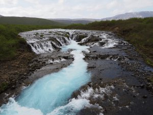 Leuchtend türkisfarbener Fluss fließt über dunkle Felsen durch eine grüne raue Landschaft in Island