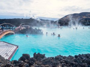 Zahlreiche Menschen entspannen im dampfenden milchblauen Wasser der Blue Lagoon zwischen dunklen Lavagesteinen in Island