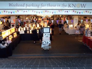 An indoor craft fair with people browsing various stalls under colorful bunting and a sign reading "Weekend parking for those in the KNOW".