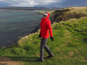 Frau in roter Jacke läuft auf einer grasbewachsenen Klippe mit Blick auf eine Bucht und entfernte Berge in Island