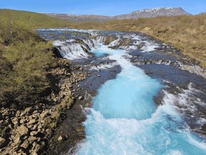 Türkisfarbener Gletscherfluss fließt durch Wasserfälle und felsige Landschaft in Island mit Bergen im Hintergrund