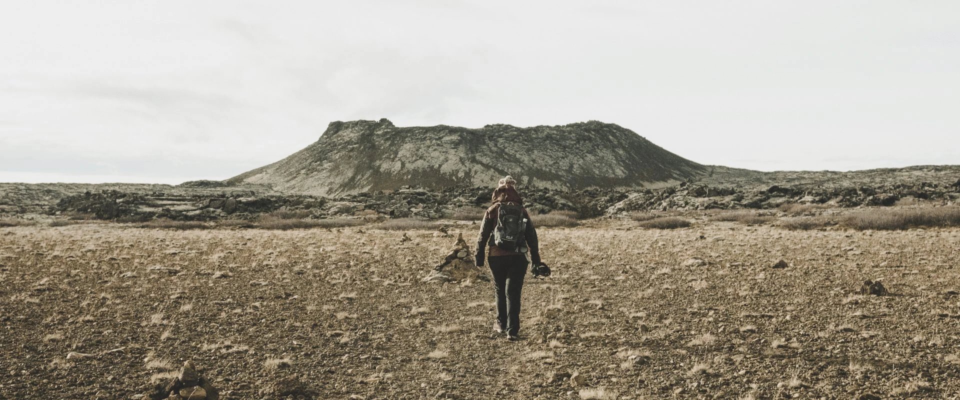 Person mit Rucksack läuft durch eine felsige karge Landschaft auf einen vulkanischen Berg in der Ferne zu in Island