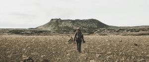 Person mit Rucksack läuft durch eine felsige karge Landschaft auf einen vulkanischen Berg in der Ferne zu in Island
