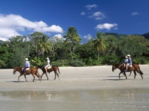 Menschen reiten an einem tropischen Strand in Australien entlang mit üppigem Wald und Bergen