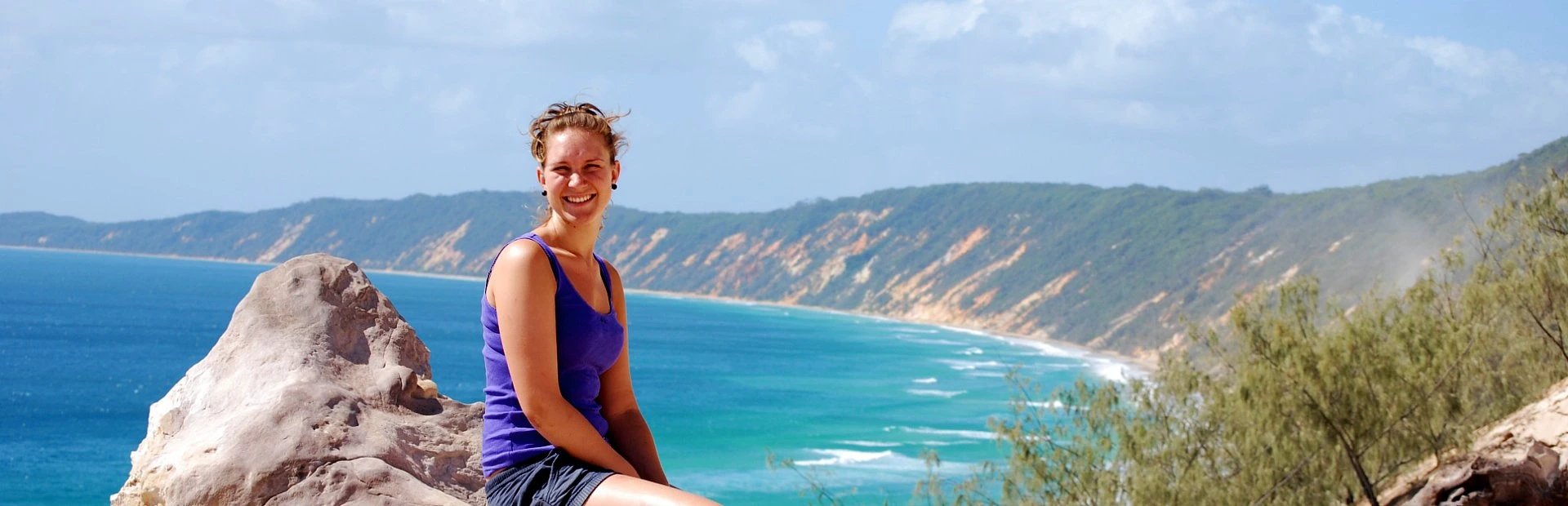 Lächelnde Frau auf einem Felsen in Australien mit Blick auf eine Küste mit türkisfarbenem Meer und farbigen Klippen