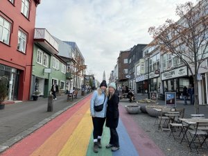 Zwei lächelnde Frauen stehen auf einer regenbogenfarbenen Straße mit bunten Häusern und der Hallgrímskirkja im Hintergrund in Reykjavík, Island