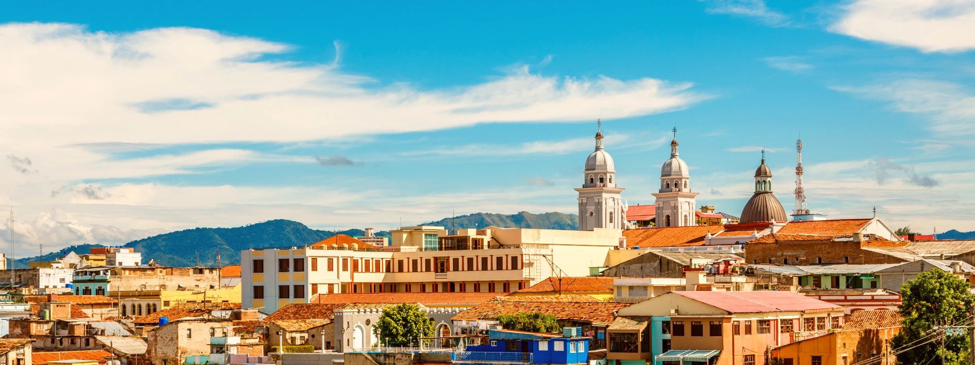 Farbenfrohes Stadtbild mit roten Dächern, zwei Kirchtürmen und Bergen unter strahlend blauem Himmel in Santiago de Cuba, Kuba