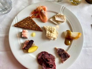 Weißer Teller mit kleinen Portionen Charcuterie, Garnelen, Fisch, dunklem Brot und Kartoffeln bei einer Mahlzeit in Grönland