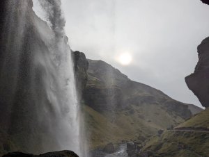 Mächtiger Wasserfall stürzt über felsige Klippen mit grüner Hügellandschaft, Wanderweg und diffuser Sonne am Himmel in Island