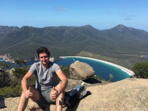 Junger Mann sitzt auf einer Felsklippe in Australien und zeigt zwei Daumen nach oben mit Blick auf eine Bucht, weißen Strand und bewaldete Berge unter blauem Himmel