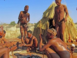 San-Menschen in traditioneller Kleidung versammeln sich um ein Feuer vor einer Strohhütte in einer trockenen Landschaft in Namibia