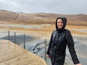 Frau in Regenjacke an einem geothermischen Gebiet mit Dampf und Schlammtümpeln in der vulkanischen Landschaft Islands