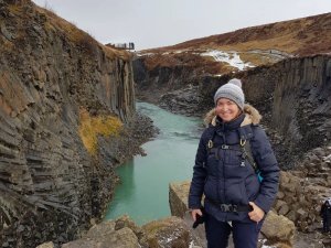 Lächelnde Frau in Winterkleidung steht vor einer Schlucht mit leuchtend türkisem Fluss und markanten Basaltsäulen in Island