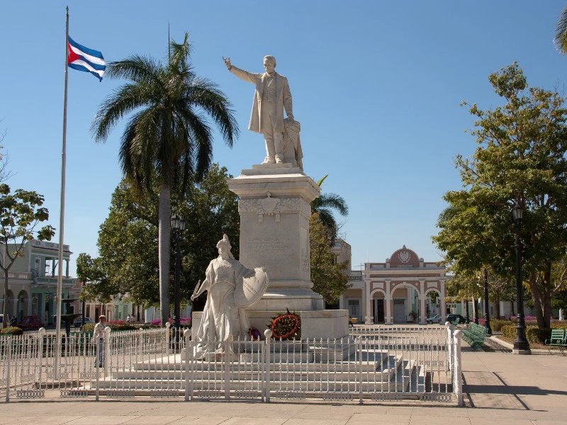 Weiße Marmorstatue eines Mannes mit ausgestrecktem Arm, wehender kubanischer Flagge und Palmen auf einem sonnigen Platz in Havanna, Kuba