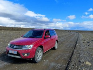 Roter SUV fährt auf einer staubigen Schotterstraße durch eine karge Landschaft unter blauem Himmel in Island