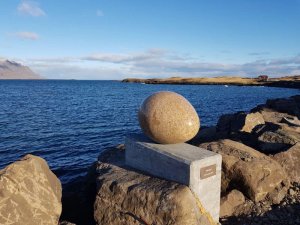 Große gesprenkelte eiförmige Steinskulptur auf grauem Sockel an einer felsigen Küste mit Blick auf das Meer und entfernte Berge in Island