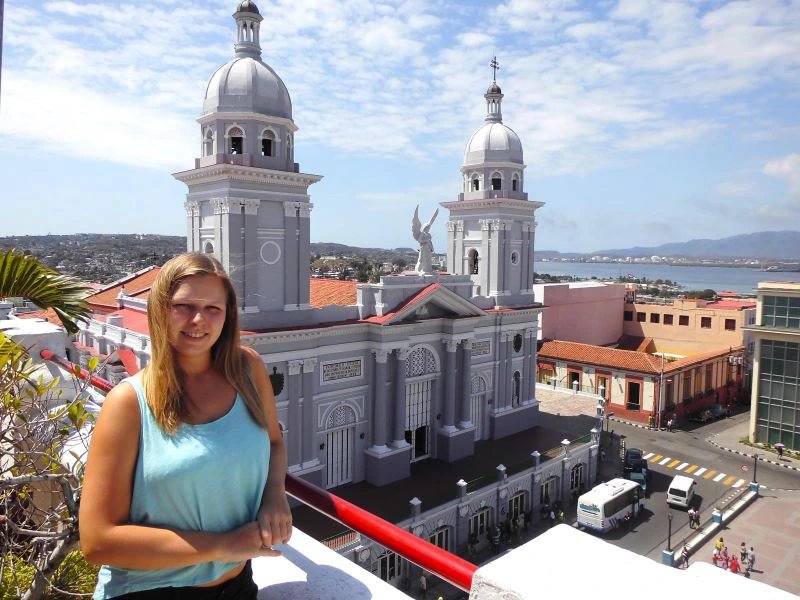Lächelnde Frau auf einem Balkon mit Blick über die Stadt, eine große graue Kathedrale mit zwei Kuppeln, eine Bucht und Berge in Santiago de Cuba, Kuba
