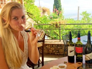 Blonde Frau trinkt Rotwein auf einer Außenterrasse mit Weinflaschen und Blick auf eine grüne Landschaft in Brda Slowenien