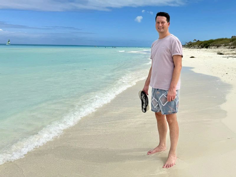 Lächelnder Mann steht barfuß am weißen Sandstrand mit türkisfarbenem Meer und Flip-Flops in der Hand in Tulum, Mexiko