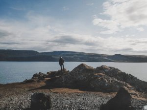 Person steht auf einer felsigen Landzunge mit Blick auf einen großen See und Berge in der Ferne unter bewölktem Himmel in Island