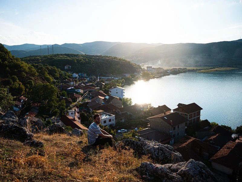Mann sitzt auf Felsen überblickt Dorf am Ohridsee bei Sonnenuntergang met bergen, Albanien