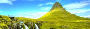 Conical green Kirkjufell mountain with a multi-tiered waterfall and a bridge in a lush landscape under a blue sky.