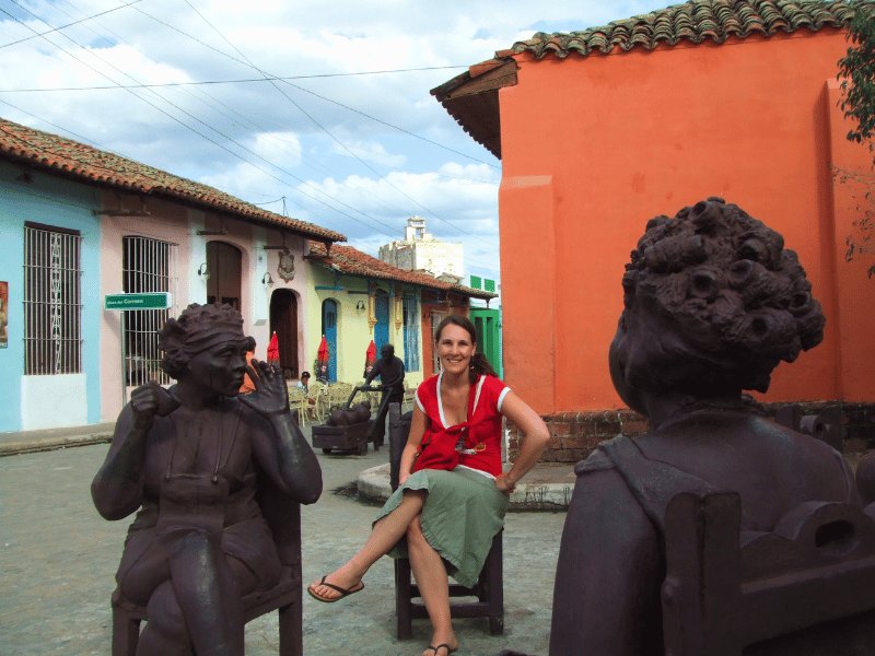 Lächelnde Frau sitzt zwischen zwei dunklen Statuen in einer farbenfrohen historischen Straße in Trinidad, Kuba