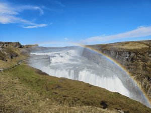 Regenbogen über einem mächtigen Wasserfall in der rauen Landschaft Islands unter klarem blauem Himmel