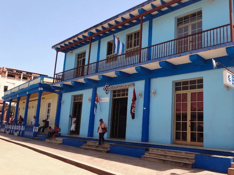 Hellblaues kubanisches Postamt mit dunkelblauen Details, Balkon und kubanischer Flagge, mit Menschen vor dem Gebäude in Baracoa, Kuba