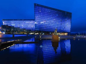 Modernes Glasgebäude leuchtet nachts blau am Wasser mit einer Statue im Vordergrund in Reykjavík, Island
